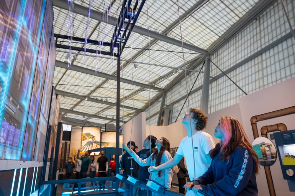 A group of teenagers pull on cords to activate an interactive water system demonstration